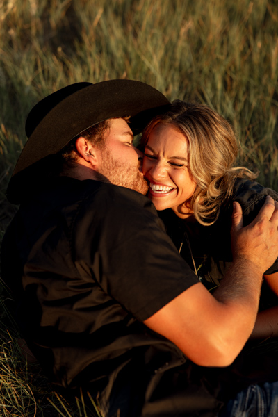 Rilee laughing while Colby kisses her cheek as they lay in the grass during their Mission Valley engagement session
