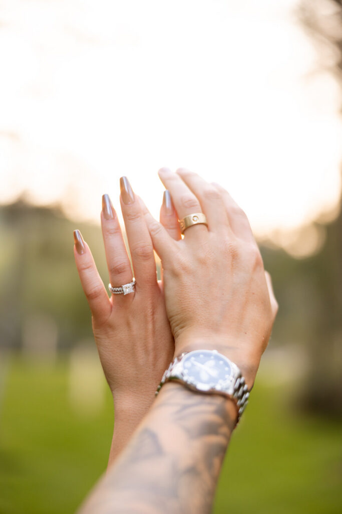 Bride and groom's wedding rings held up against golden hour light, his tattooed wrist and silver watch visible below — Mikayla + Cayden — Haymoon Resort Wedding — Whitefish Montana — Stacy Edwards Photography