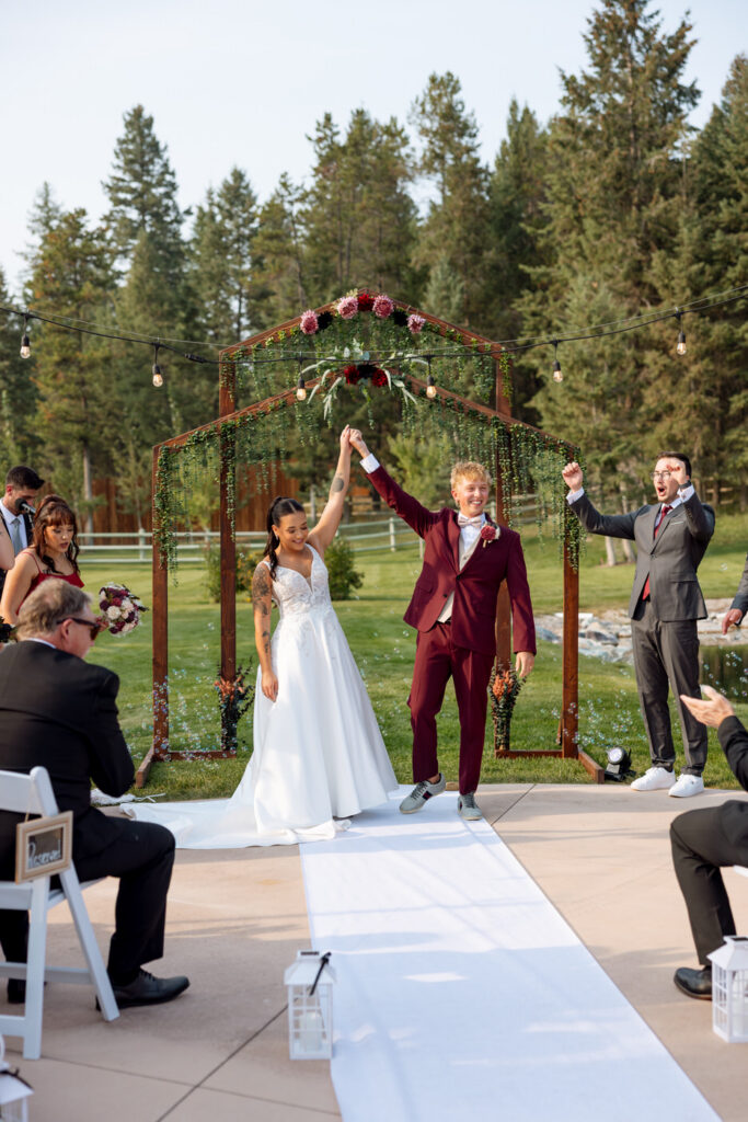Bride and groom raising their hands in celebration as they walk back down the aisle after ceremony beneath a wooden floral arch, guests cheering — Mikayla + Cayden — Haymoon Resort Wedding — Whitefish Montana — Stacy Edwards Photography