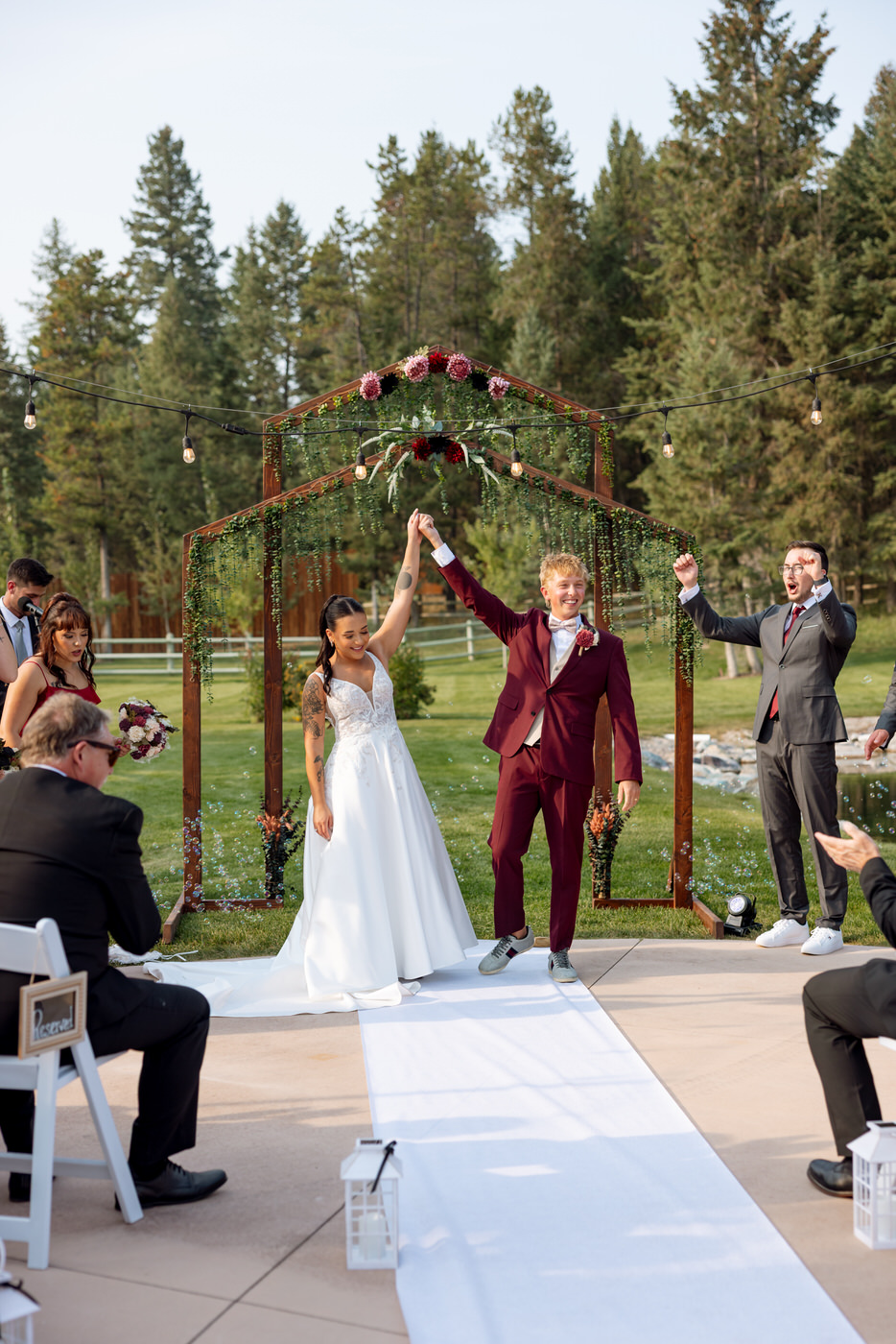 Bride and groom raising their hands in celebration as they walk back down the aisle after ceremony beneath a wooden floral arch, guests cheering — Mikayla + Cayden — Haymoon Resort Wedding — Whitefish Montana — Stacy Edwards Photography