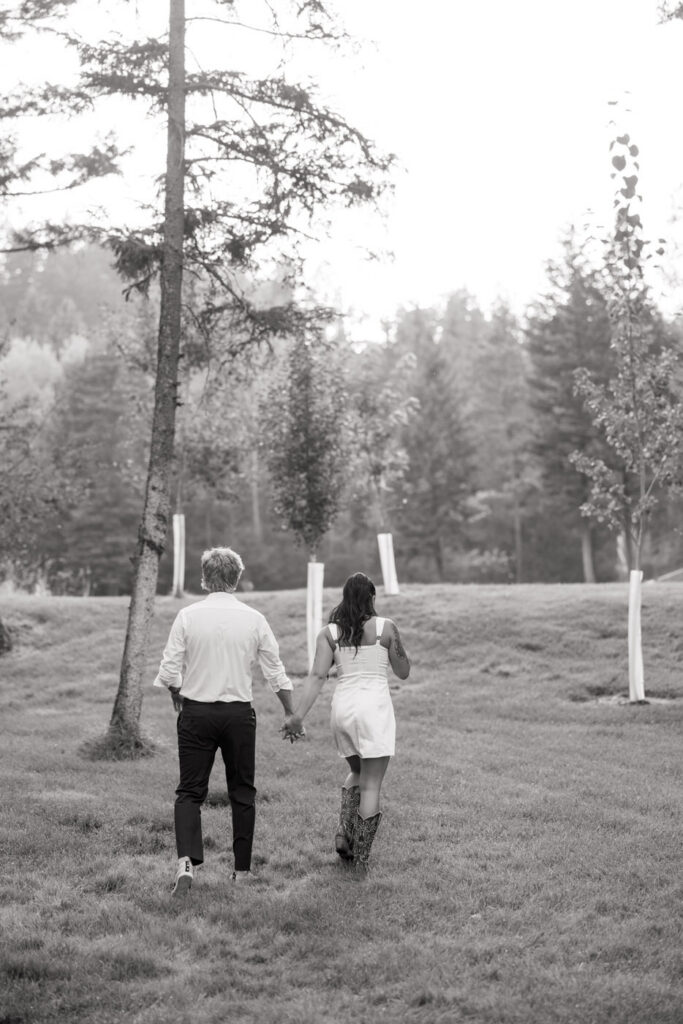 Black and white photo of bride and groom walking hand in hand away from camera across a sweeping Montana lawn with pine trees — Mikayla + Cayden — Haymoon Resort Wedding — Whitefish Montana — Stacy Edwards Photography