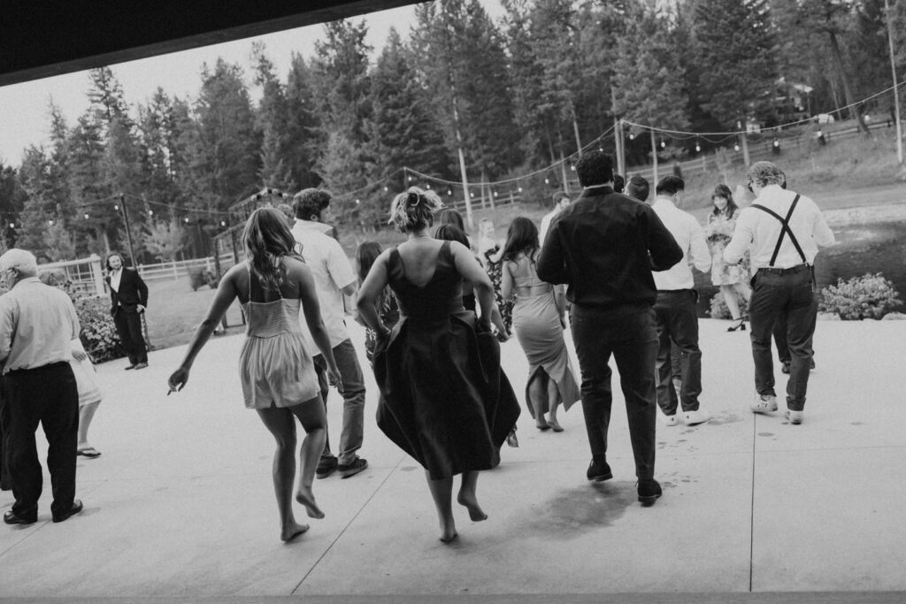 Black and white wide shot of wedding guests flooding the outdoor dance floor at dusk, string lights overhead and Montana pines in the background — Mikayla + Cayden — Haymoon Resort Wedding — Whitefish Montana — Stacy Edwards Photography