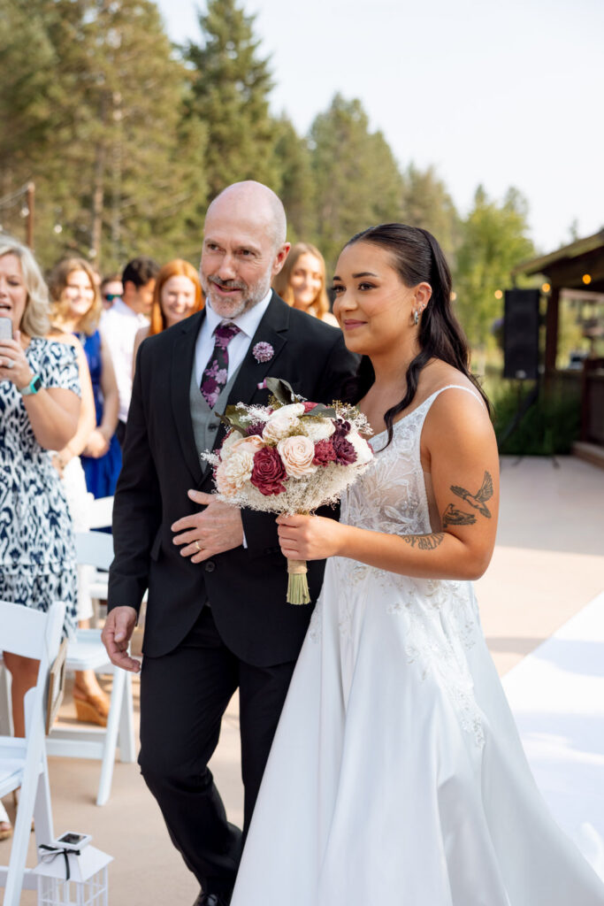 Bride walking down the aisle on the arm of her father, holding a burgundy and blush bouquet, guests watching from white chairs — Mikayla + Cayden — Haymoon Resort Wedding — Whitefish Montana — Stacy Edwards Photography