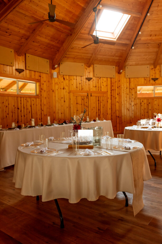 Haymoon Resort reception hall interior with warm wood-paneled walls, vaulted ceiling with skylights, and round tables set with cream linens and burgundy florals — Mikayla + Cayden — Haymoon Resort Wedding — Whitefish Montana — Stacy Edwards Photography