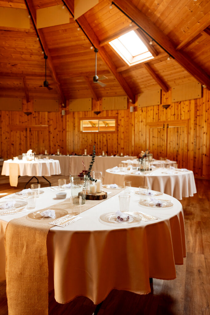 Haymoon Resort reception hall set for dinner with warm wood ceiling, skylights, round tables draped in cream and burlap runners, and candlelit centerpieces — Mikayla + Cayden — Haymoon Resort Wedding — Whitefish Montana — Stacy Edwards Photography