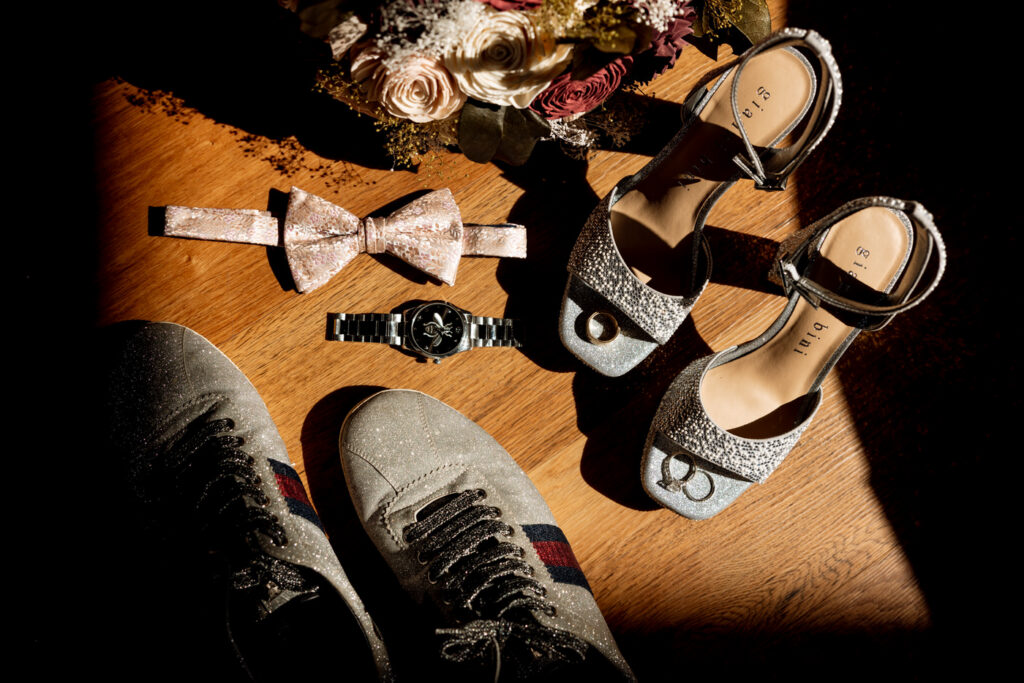 Flat lay of wedding day details including sparkly bridal heels, groom's sneakers, blush bow tie, silver watch, wedding rings, and burgundy bouquet arranged on hardwood floor — Mikayla + Cayden — Haymoon Resort Wedding — Whitefish Montana — Stacy Edwards Photography
