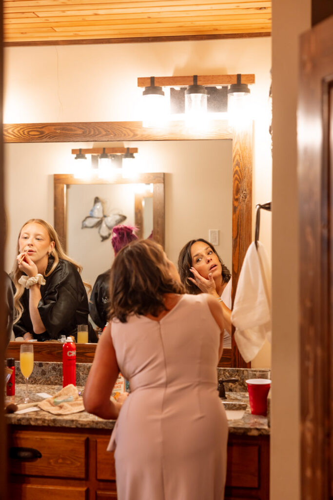 Bridesmaids doing hair and makeup together in the Haymoon Resort cabin bathroom mirror, champagne and hair products on the counter — Mikayla + Cayden — Haymoon Resort Wedding — Whitefish Montana — Stacy Edwards Photography