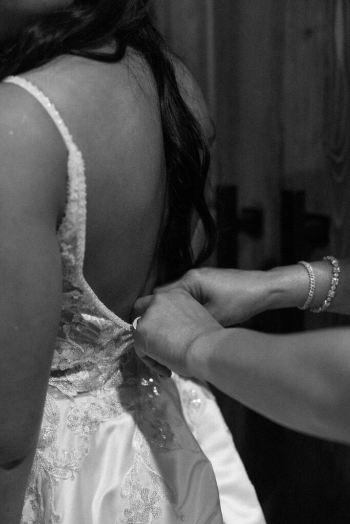 Black and white close-up of hands buttoning the back of the bride's lace wedding dress during getting ready — Mikayla + Cayden — Haymoon Resort Wedding — Whitefish Montana — Stacy Edwards Photography