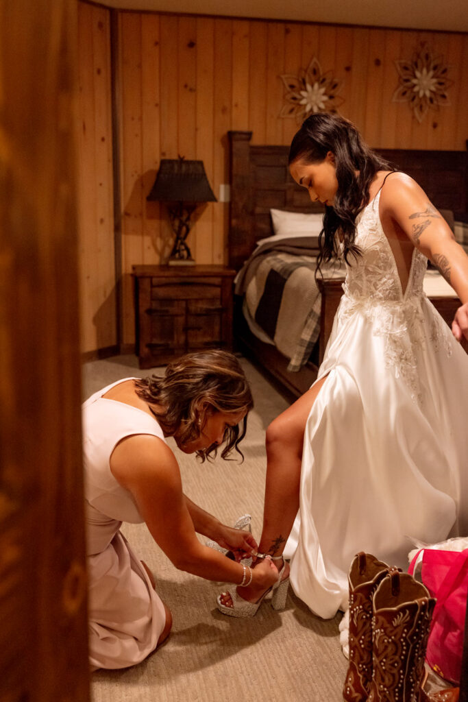 Brides mom kneeling to buckle the bride's crystal platform heels in a warm wood-paneled Haymoon Resort cabin room, cowboy boots visible nearby — Mikayla + Cayden — Haymoon Resort Wedding — Whitefish Montana — Stacy Edwards Photography