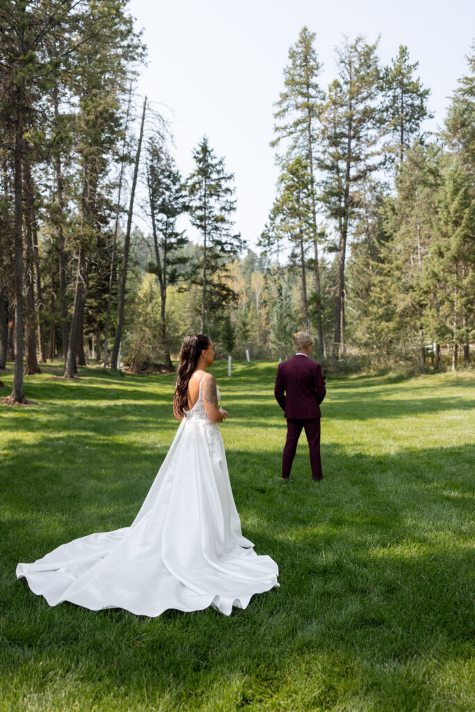 Bride approaching groom from behind for their first look on the green lawn at Haymoon Resort, her cathedral train spread behind her and Montana pines framing the scene — Mikayla + Cayden — Haymoon Resort Wedding — Whitefish Montana — Stacy Edwards Photography