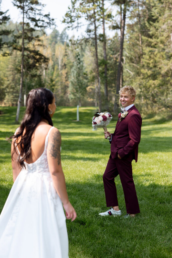 Groom turning around during first look to see his bride, beaming in his burgundy suit while holding her bouquet, pine trees and green grass behind him — Mikayla + Cayden — Haymoon Resort Wedding — Whitefish Montana — Stacy Edwards Photography