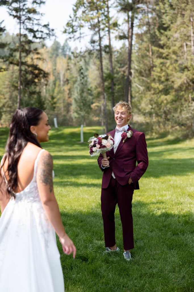 Groom laughing with joy at his first look with the bride on the Haymoon Resort lawn, holding her bouquet of burgundy and blush flowers — Mikayla + Cayden — Haymoon Resort Wedding — Whitefish Montana — Stacy Edwards Photography