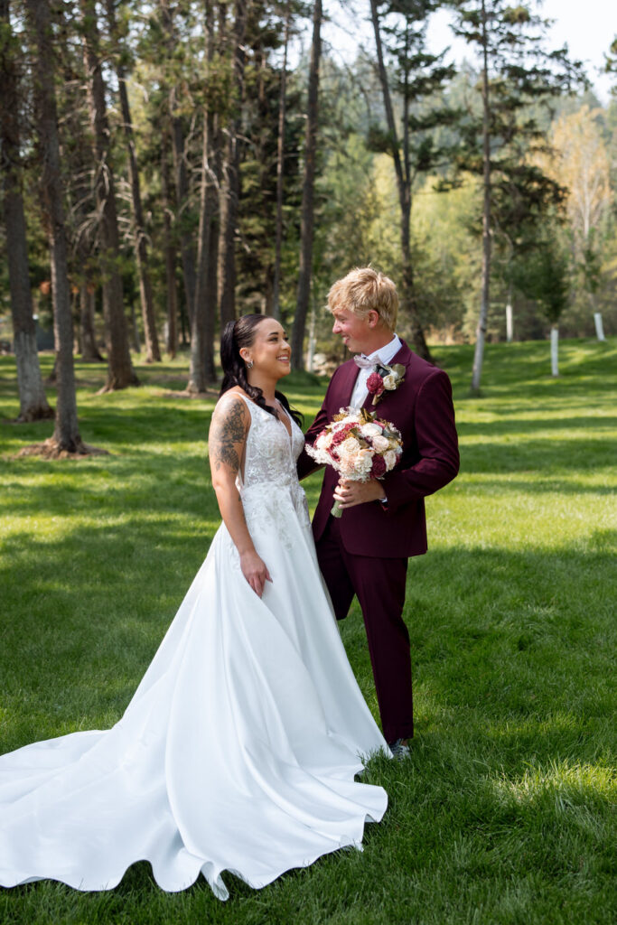 Bride and groom smiling at each other on the Haymoon Resort lawn, her lace dress train spreading across the grass and tall pines filling the background — Mikayla + Cayden — Haymoon Resort Wedding — Whitefish Montana — Stacy Edwards Photography