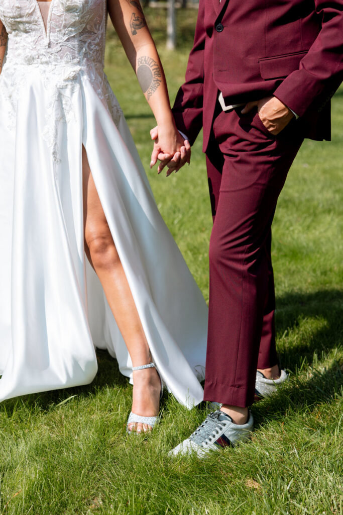 Close-up of bride and groom holding hands, her crystal heels and floral arm tattoo visible, his burgundy suit pants and designer sneakers shown — Mikayla + Cayden — Haymoon Resort Wedding — Whitefish Montana — Stacy Edwards Photography