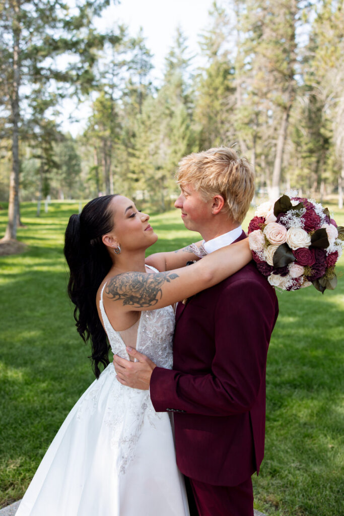 Bride looking up at groom during couples portraits, arms around each other, her back-detail lace gown and floral tattoo visible against green Montana lawn — Mikayla + Cayden — Haymoon Resort Wedding — Whitefish Montana — Stacy Edwards Photography
