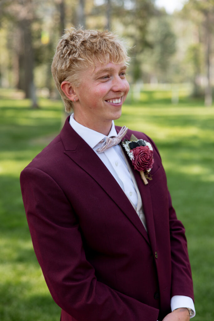 Groom solo portrait in burgundy suit and blush bow tie with rose boutonnière, smiling off to the side against a Montana pine tree backdrop — Mikayla + Cayden — Haymoon Resort Wedding — Whitefish Montana — Stacy Edwards Photography