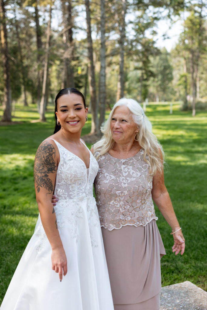 Bride and older woman smiling together for a family portrait on the Haymoon Resort lawn, both looking at the camera against a backdrop of tall pines — Mikayla + Cayden — Haymoon Resort Wedding — Whitefish Montana — Stacy Edwards Photography