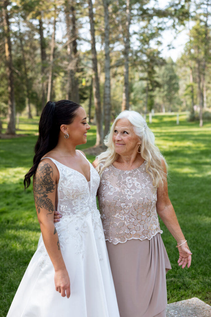 Bride and older woman sharing a candid laugh together outdoors on the green lawn at Haymoon Resort, pine trees behind them — Mikayla + Cayden — Haymoon Resort Wedding — Whitefish Montana — Stacy Edwards Photography