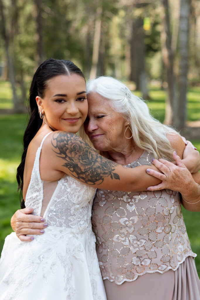 Bride and older woman hugging warmly in an outdoor portrait, bride's floral sleeve tattoo visible, pine trees and green grass behind them — Mikayla + Cayden — Haymoon Resort Wedding — Whitefish Montana — Stacy Edwards Photography