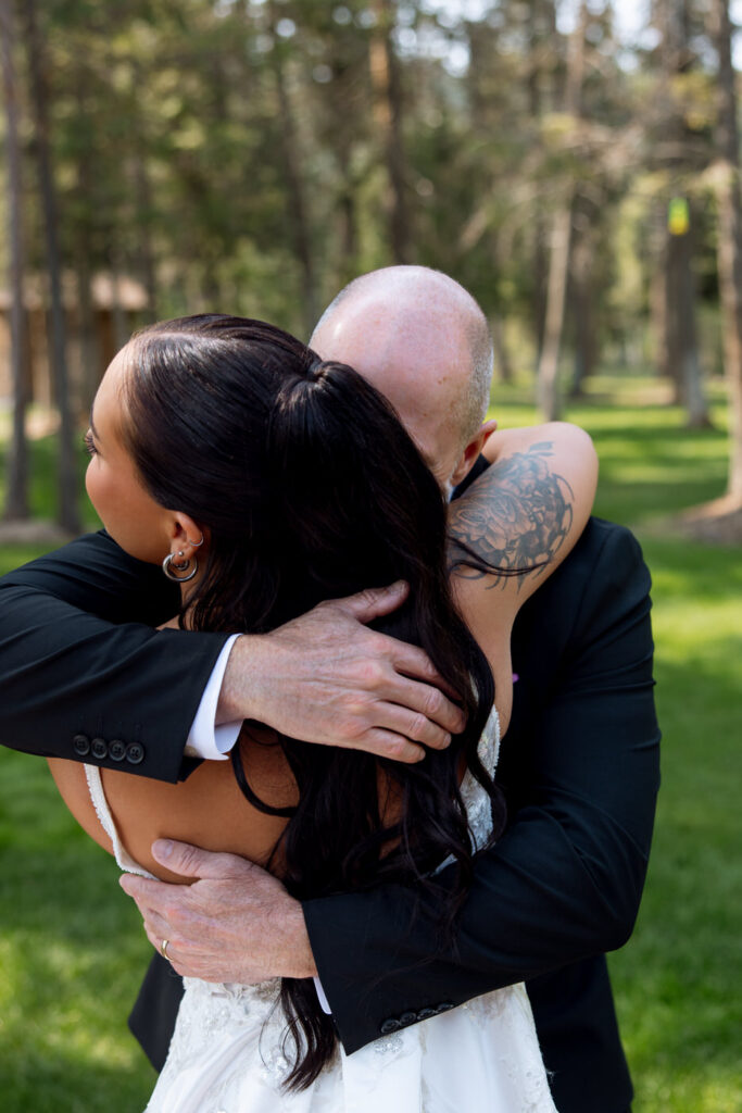 Bride being embraced by an older man in a black suit in a tight, emotional hug on the Haymoon Resort lawn, pine trees in the background — Mikayla + Cayden — Haymoon Resort Wedding — Whitefish Montana — Stacy Edwards Photography