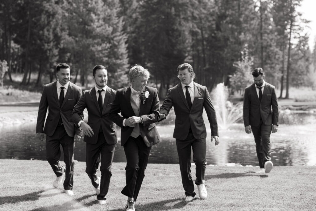 Black and white photo of groom and groomsmen walking confidently across the Haymoon Resort lawn past the pond and fountain, laughing together in dark suits — Mikayla + Cayden — Haymoon Resort Wedding — Whitefish Montana — Stacy Edwards Photography