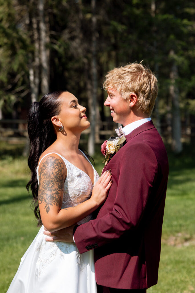 Bride and groom sharing a tender moment in front of pine trees, she in lace wedding dress with floral sleeve tattoo, he in burgundy suit with boutonnière — Mikayla + Cayden — Haymoon Resort Wedding — Whitefish Montana — Stacy Edwards Photography