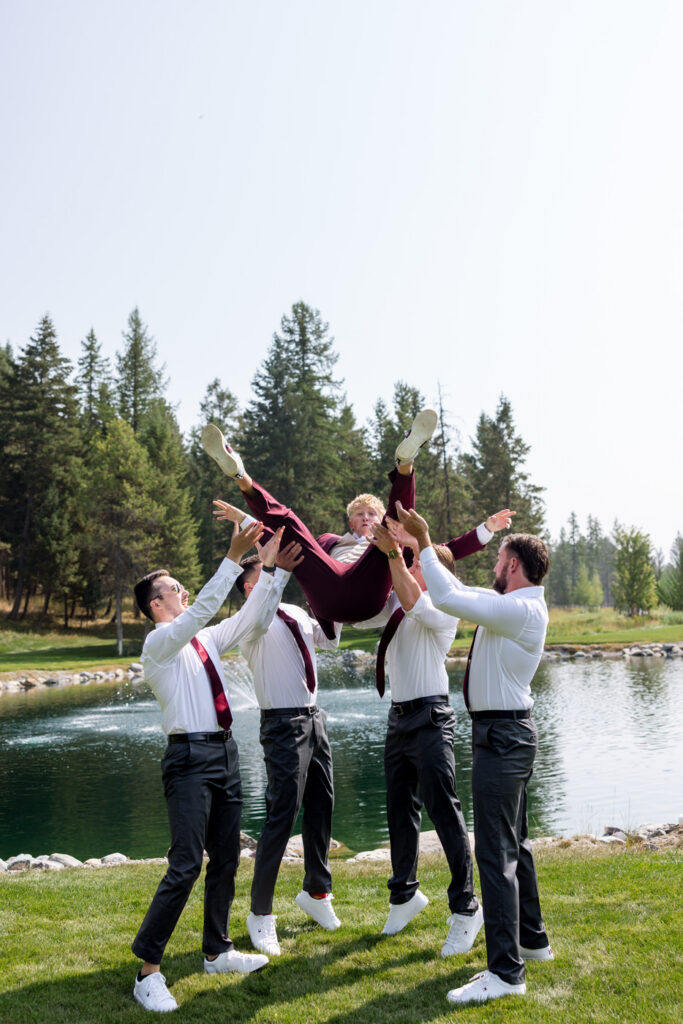 Groomsmen tossing the groom into the air in front of the Haymoon Resort pond and fountain, everyone laughing in white shirts and burgundy ties — Mikayla + Cayden — Haymoon Resort Wedding — Whitefish Montana — Stacy Edwards Photography