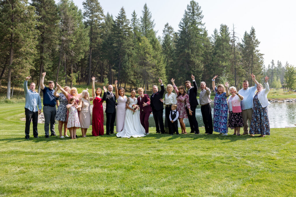 Large wedding family group photo on the Haymoon Resort lawn with everyone raising their hands in celebration, pond and Montana pine trees behind them — Mikayla + Cayden — Haymoon Resort Wedding — Whitefish Montana — Stacy Edwards Photography