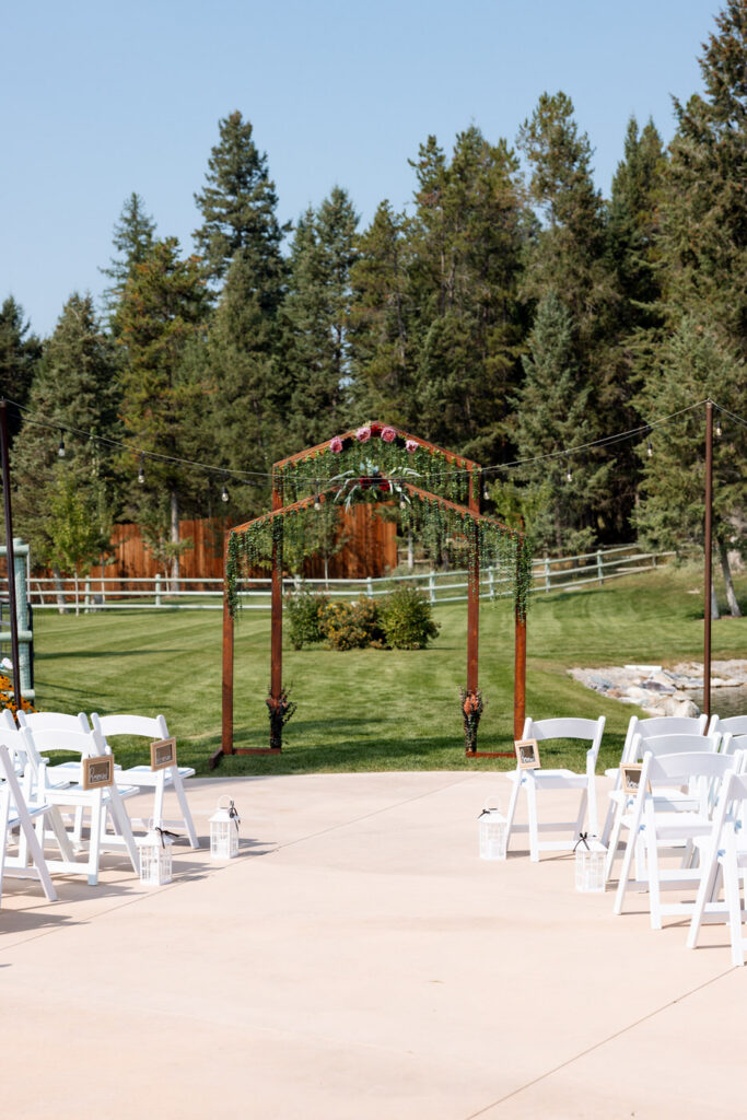 Wide shot of the Haymoon Resort outdoor ceremony space showing the wooden arch draped in greenery and burgundy florals, white chairs lining the aisle, and Montana pine trees beyond — Mikayla + Cayden — Haymoon Resort Wedding — Whitefish Montana — Stacy Edwards Photography
