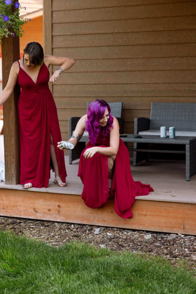 Candid of bridesmaid with purple hair crouching on the cabin porch steps laughing while holding a drink, the other bridesmaid in burgundy standing beside her — Mikayla + Cayden — Haymoon Resort Wedding — Whitefish Montana — Stacy Edwards Photography