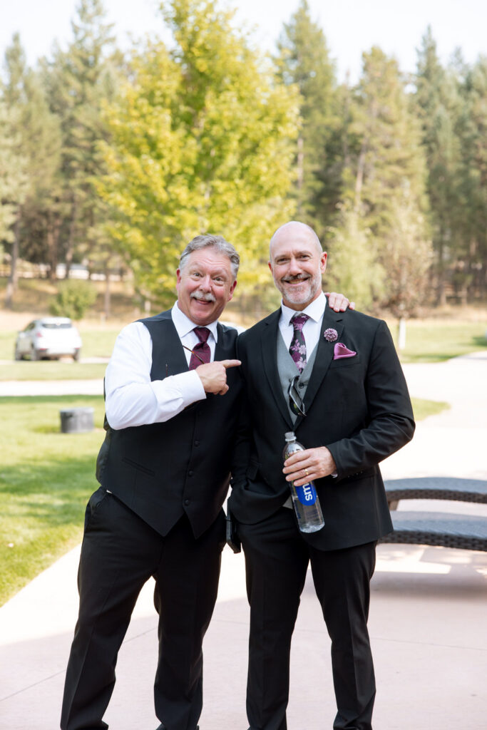 Two fathers of the wedding party smiling and joking together outdoors at Haymoon Resort before the ceremony, both in dark suits with burgundy ties — Mikayla + Cayden — Haymoon Resort Wedding — Whitefish Montana — Stacy Edwards Photography
