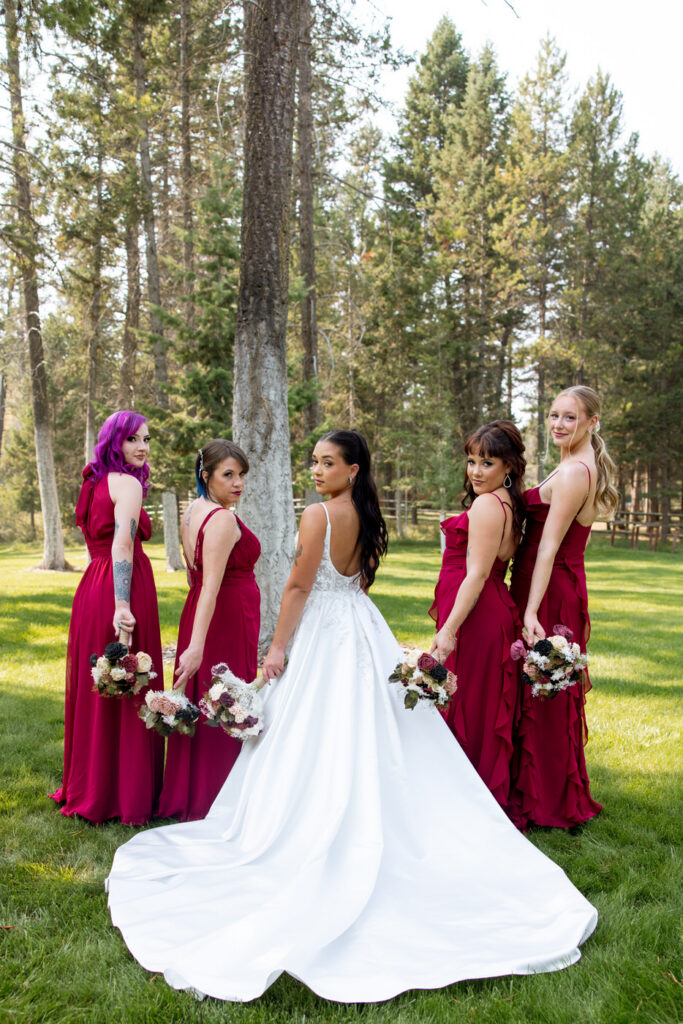 Bride and four bridesmaids in burgundy dresses posed looking back over their shoulders on the Montana lawn at Haymoon Resort, bride's cathedral train spread across the grass — Mikayla + Cayden — Haymoon Resort Wedding — Whitefish Montana — Stacy Edwards Photography