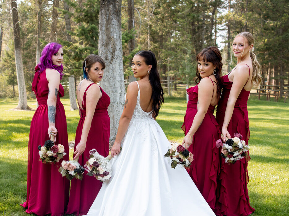 Bride and four bridesmaids in burgundy dresses posed looking back over their shoulders on the Montana lawn at Haymoon Resort, bride's cathedral train spread across the grass — Mikayla + Cayden — Haymoon Resort Wedding — Whitefish Montana — Stacy Edwards Photography