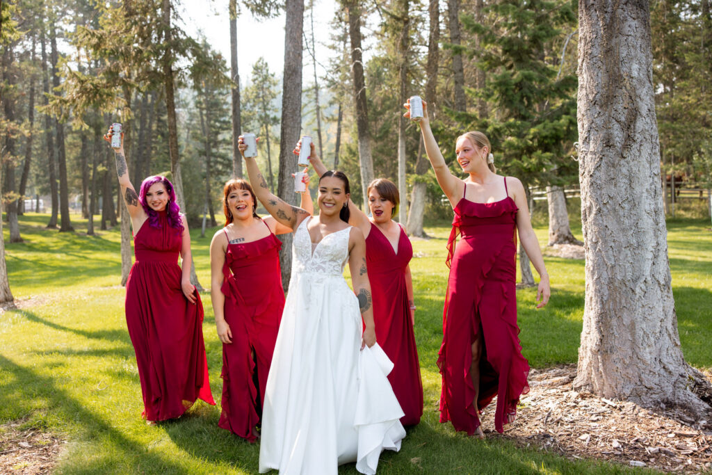 Bride and bridesmaids in burgundy dresses walking through the pine trees at Haymoon Resort raising drinks in celebration and laughing — Mikayla + Cayden — Haymoon Resort Wedding — Whitefish Montana — Stacy Edwards Photography