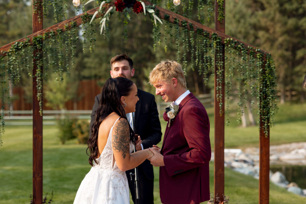 Bride and groom laughing together during outdoor ceremony beneath a wooden arch draped in greenery and burgundy florals, officiant visible behind them — Mikayla + Cayden — Haymoon Resort Wedding — Whitefish Montana — Stacy Edwards Photography