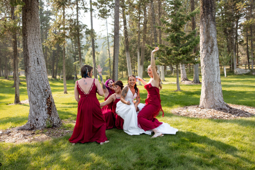Bride and bridesmaids in burgundy dresses sitting together on the Montana lawn in the trees, laughing and raising their arms, a joyful candid moment — Mikayla + Cayden — Haymoon Resort Wedding — Whitefish Montana — Stacy Edwards Photography
