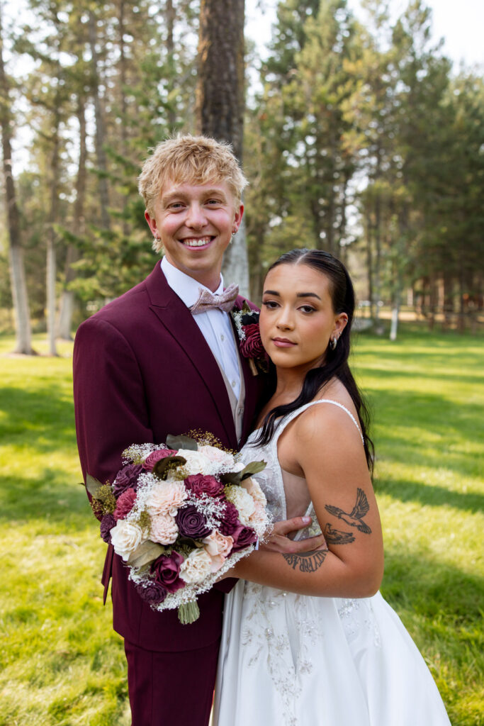 Bride and groom close portrait facing camera, he smiling broadly in burgundy suit, she resting against him holding the bouquet, pine trees behind them — Mikayla + Cayden — Haymoon Resort Wedding — Whitefish Montana — Stacy Edwards Photography