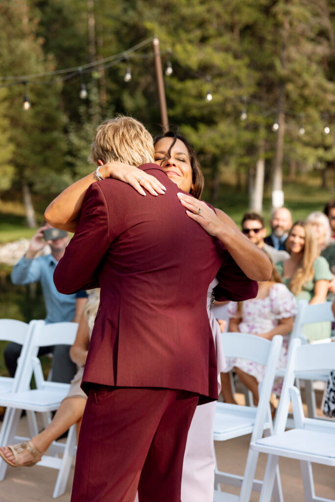 Groom being embraced by a family member at the ceremony altar, guests seated in white chairs visible behind them, string lights and pine trees in the background — Mikayla + Cayden — Haymoon Resort Wedding — Whitefish Montana — Stacy Edwards Photography