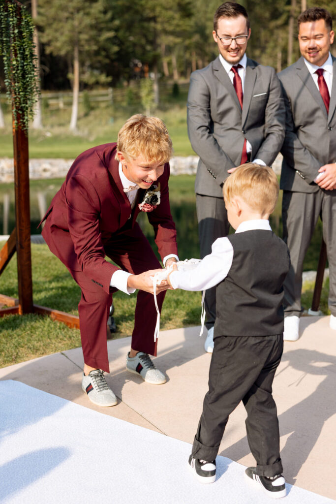 Groom bending down to receive the ring pillow from the ring bearer at the Haymoon Resort ceremony altar, groomsmen smiling behind them — Mikayla + Cayden — Haymoon Resort Wedding — Whitefish Montana — Stacy Edwards Photography