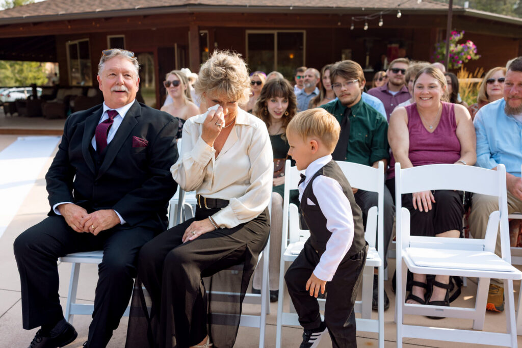 Candid of a woman in the front row dabbing tears at the ceremony while a young ring bearer in a tiny vest and bow tie wanders beside her, guests watching behind — Mikayla + Cayden — Haymoon Resort Wedding — Whitefish Montana — Stacy Edwards Photography