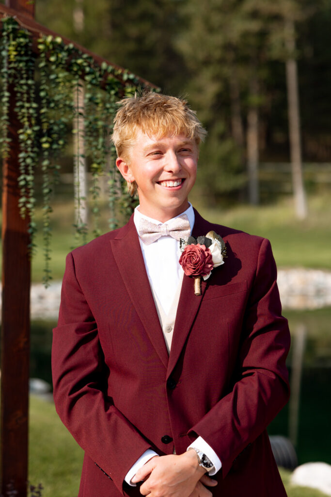 Groom beaming at the ceremony altar beside the wooden arch draped in greenery, Haymoon Resort pond visible in the soft background — Mikayla + Cayden — Haymoon Resort Wedding — Whitefish Montana — Stacy Edwards Photography