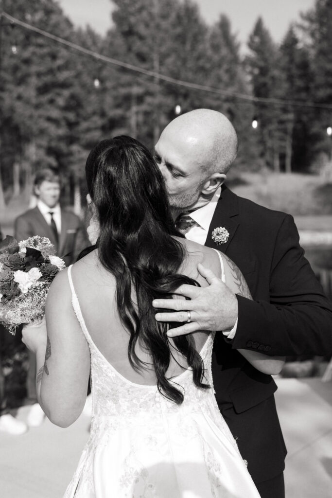 Black and white photo of father kissing the bride's cheek at the ceremony altar as the groom watches in the background, string lights overhead — Mikayla + Cayden — Haymoon Resort Wedding — Whitefish Montana — Stacy Edwards Photography