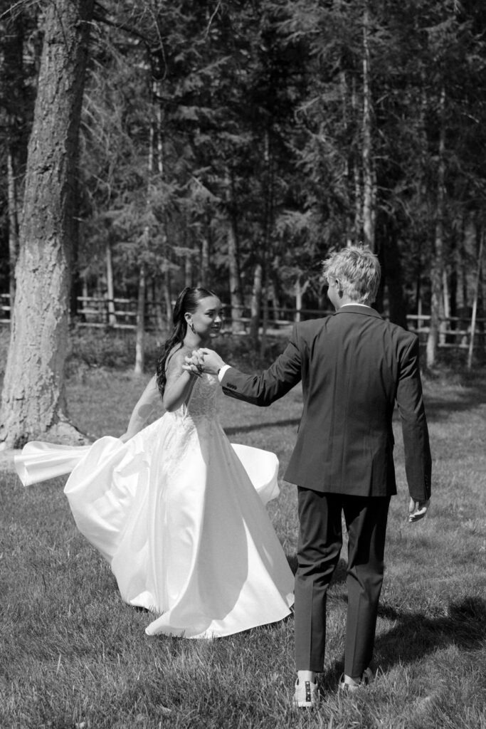 Black and white photo of groom spinning bride on a Montana lawn surrounded by tall pine trees, her dress billowing — Mikayla + Cayden — Haymoon Resort Wedding — Whitefish Montana — Stacy Edwards Photography