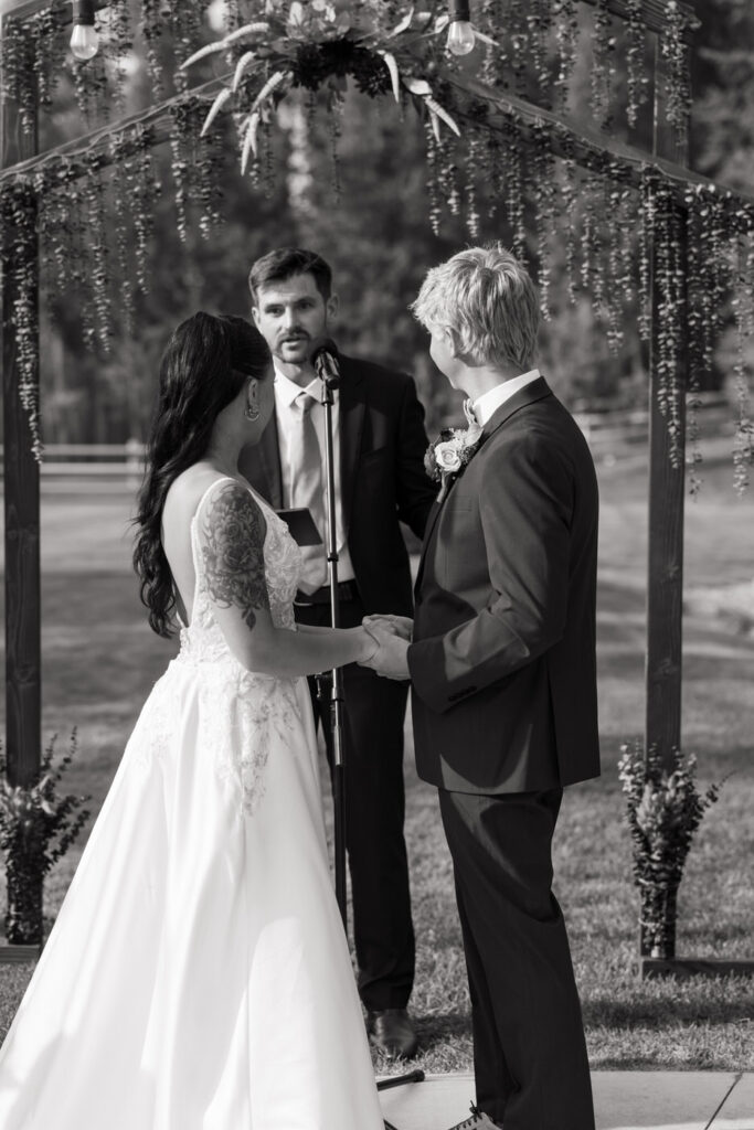 Black and white ceremony photo of bride and groom holding hands and facing each other beneath the greenery-draped wooden arch while the officiant speaks — Mikayla + Cayden — Haymoon Resort Wedding — Whitefish Montana — Stacy Edwards Photography
