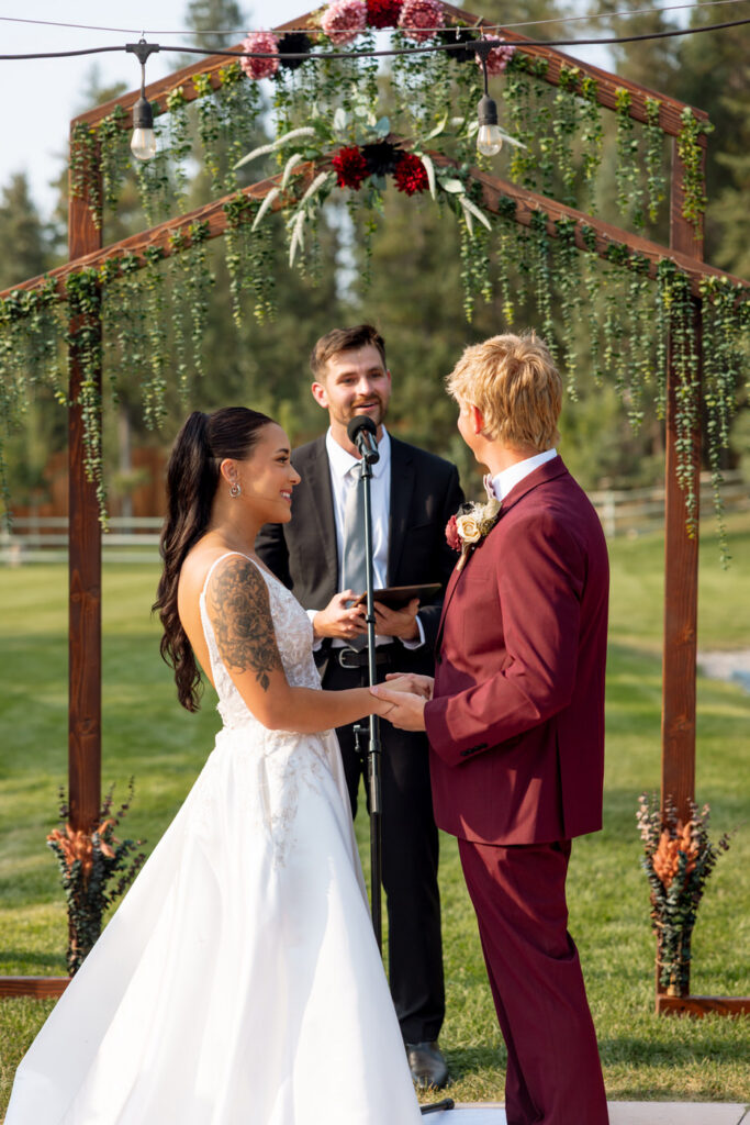 Bride smiling at groom during outdoor ceremony beneath the Haymoon Resort wooden arch draped in cascading greenery and burgundy florals, officiant behind them — Mikayla + Cayden — Haymoon Resort Wedding — Whitefish Montana — Stacy Edwards Photography