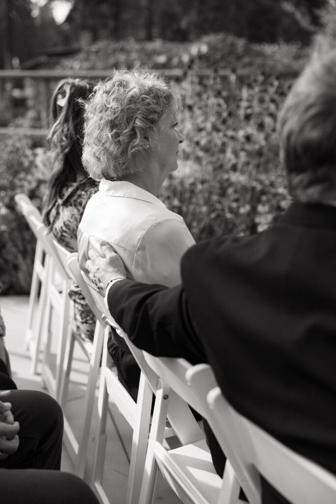 Black and white candid of an older woman seated in a white ceremony chair with a hand placed gently on her back by the person beside her — Mikayla + Cayden — Haymoon Resort Wedding — Whitefish Montana — Stacy Edwards Photography