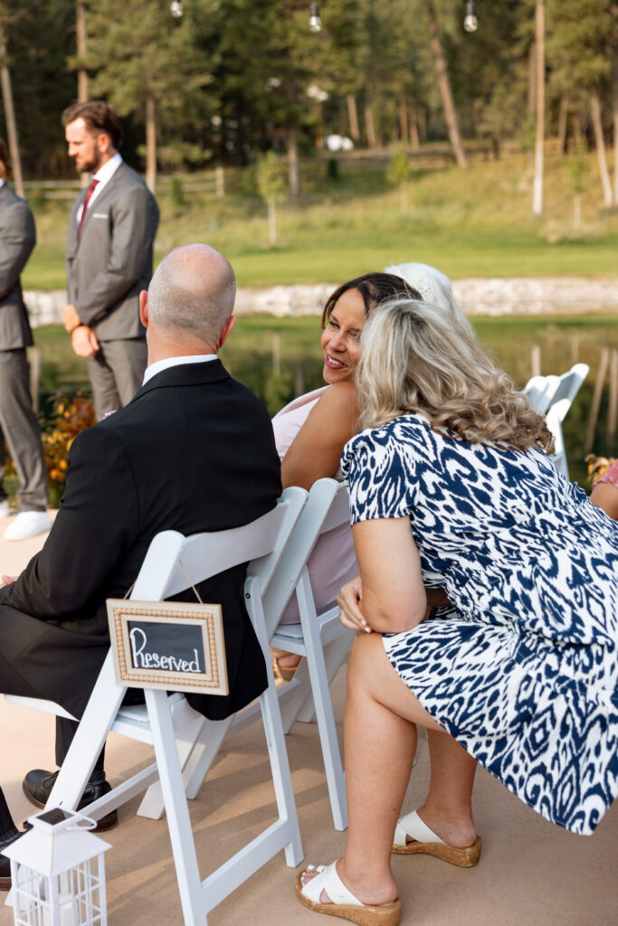 Candid of family members seated in reserved front-row ceremony chairs sharing a warm moment before the Haymoon Resort wedding ceremony begins, pond in the background — Mikayla + Cayden — Haymoon Resort Wedding — Whitefish Montana — Stacy Edwards Photography