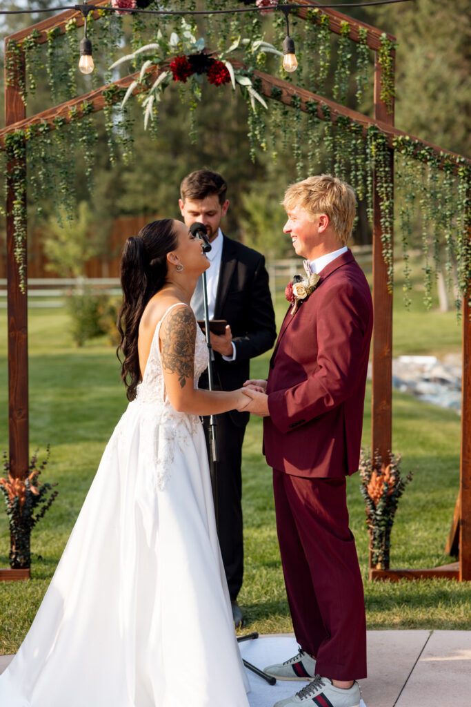 Bride laughing and looking up at groom during the outdoor ceremony beneath the greenery-draped wooden arch at Haymoon Resort, both holding hands — Mikayla + Cayden — Haymoon Resort Wedding — Whitefish Montana — Stacy Edwards Photography