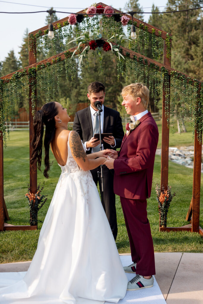 Bride and groom laughing together at the altar beneath the wooden arch at Haymoon Resort as the officiant reads, Flathead Valley pine trees glowing in background — Mikayla + Cayden — Haymoon Resort Wedding — Whitefish Montana — Stacy Edwards Photography