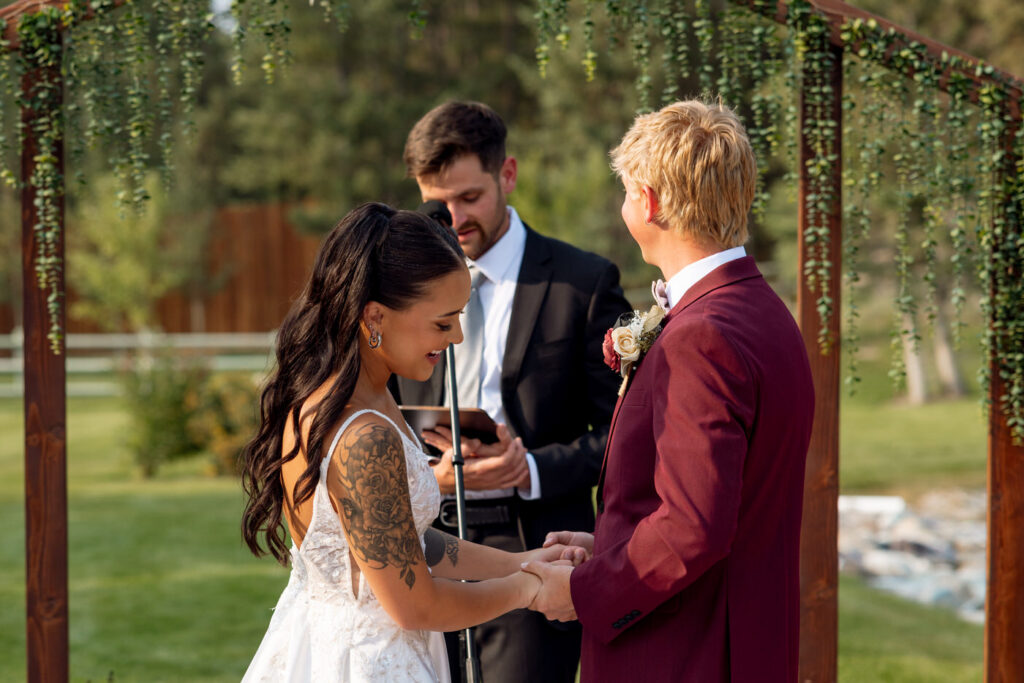 Bride speaking her vows at the altar beneath the greenery arch at Haymoon Resort while groom listens and smiles, pond and white fence in the background — Mikayla + Cayden — Haymoon Resort Wedding — Whitefish Montana — Stacy Edwards Photography